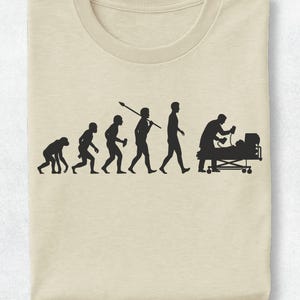 Può includere: Una t-shirt color beige con una stampa grafica nera che raffigura l'evoluzione dell'umanità, partendo da un primate e progredendo attraverso varie fasi dello sviluppo umano, terminando con una persona su un letto d'ospedale che riceve cure.