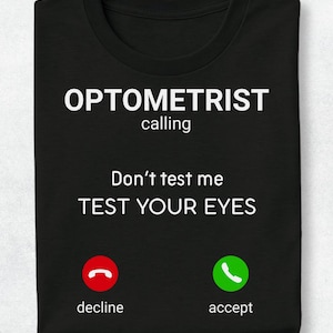 May include: A black t-shirt featuring white text that reads OPTOMETRIST calling, Don't test me, TEST YOUR EYES. Below the text are two circular icons: a red phone icon labelled decline and a green phone icon labelled accept, mimicking a call screen.