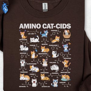 Könnte beinhalten: Ein dunkelbraunes Sweatshirt mit Rundhalsausschnitt und einem Grafikdruck mit dem Titel AMINO CAT-CIDS. Das Design zeigt verschiedene Cartoon-Katzen, gepaart mit chemischen Strukturen und Wortspiel-Namen.