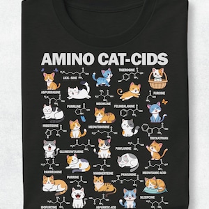 Op de afbeelding: Een zwart t-shirt met de tekst AMINO CAT-CIDS bovenaan. De afbeelding toont verschillende cartoonkatten gecombineerd met chemische structuren en op woordspelingen gebaseerde namen zoals Lick-sine, Purrline, Meowthionine, Pawreoning en Sleepcine.