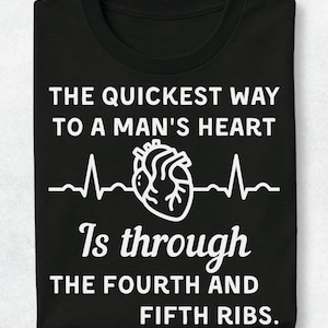 Op de afbeelding: Een zwart t-shirt met witte tekst die luidt THE QUICKEST WAY TO A MAN'S HEART Is through THE FOURTH AND FIFTH RIBS. Een witte lijntekening van een menselijk hart met een ECG-hartslaglijn is in het midden van het ontwerp geplaatst.