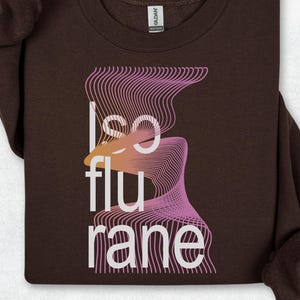 May include: A dark brown crewneck sweatshirt featuring the white text "isoflurane" printed vertically. The design includes a graphic element of wavy, thin lines in shades of pink and orange that intersect with the letters of the word.