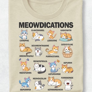 Könnte beinhalten: Ein beiges T-Shirt mit dem Text MEOWDICATIONS oben, gefolgt von einem Raster aus fünfzehn Cartoon-Katzenillustrationen, die jeweils mit einem wortspielbasierten Namen wie Clawpidogrel, Catropine, Lidocatine, Gabapawtin und anderen in Schwarz versehen sind.