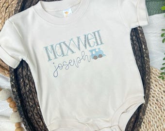 May include: Cream-colored baby bodysuit with the name "Maxwell Joseph" embroidered in blue thread, along with a small train graphic. The bodysuit has short sleeves and snap closures.