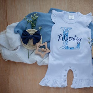 May include: White baby onesie with a blue floral letter L and the name "Liberty" embroidered on the front. The onesie has ruffled sleeves and a ruffled bottom.