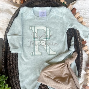 May include: A light blue baby onesie with a green and white plaid letter R embroidered on the front. The onesie has the name "Ryder McClain" embroidered below the letter. A brown knit hat is included.