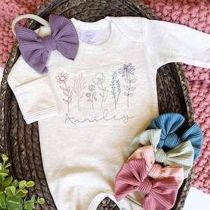 May include: A beige baby onesie with a floral embroidery design and the name "Annsiley" embroidered below. The onesie is displayed on a woven basket with a purple and a set of four multi-colored hair bows.