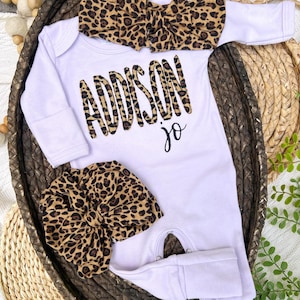 Puede incluir: Un conjunto para bebé blanco con el nombre "Addison Jo" en estampado de leopardo, acompañado de una diadema a juego y un gorro anudado. El conjunto se muestra en una cesta tejida, mostrando un atuendo coordinado para bebé.
