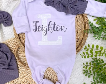 Può includere: Un set di abbigliamento per neonati color lavanda. Il set include una tutina a maniche lunghe con il nome "Leighton" e una grande "L". Sono inclusi anche una fascia abbinata e un cappello in stile turbante in un colore grigio-viola coordinato.