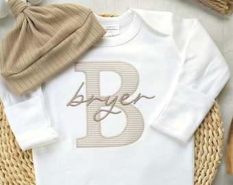 Könnte beinhalten: Weißer Baby-Strampler mit langen Ärmeln und einem braunen gestickten Buchstaben B mit dem Namen "Bryer" darunter. Der Strampler liegt auf einer geflochtenen Strohhütte mit einer braunen Strickmütze und Holzspielzeug.