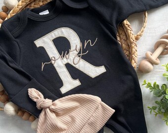 May include: Black baby onesie with a large, off-white letter "R" and the name "Rowyn" embroidered in a delicate script. A matching ribbed, light pink knotted hat is included. The onesie has long sleeves and footed bottoms.