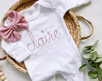 May include: White baby onesie with long sleeves and a pink embroidered name "claire" on the front. The onesie is lying on a woven basket with a pink bow headband and a wooden bead garland.