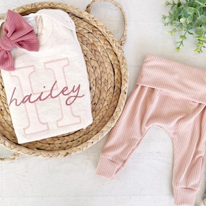 May include: A baby outfit set featuring a cream coloured onesie with the letter "H" and the name "hailey" embroidered in pink. The set also includes a pair of pink ribbed trousers.