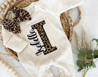 May include: A beige baby onesie with long sleeves and a snap closure. The onesie has a large letter "I" made of a leopard print fabric and the word "Hello" written in black script. A leopard print bow is on the onesie.