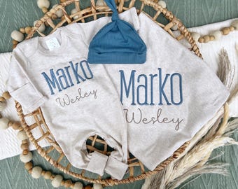 May include: A beige baby outfit set with the name "Marko" embroidered in blue, and "Wesley" in gray. Includes a matching blue ribbed knit hat. The set is displayed on a woven basket.