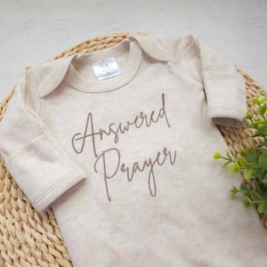 Op de afbeelding: Een beige baby-romper met lange mouwen met de tekst "Answered Prayer" geborduurd in bruin.