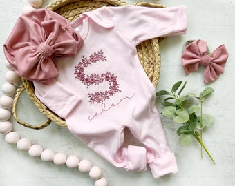 Puede incluir: Un body de bebé de color rosa claro de manga larga con una letra S floral bordada en hilo rosa oscuro, con el nombre Salem bordado debajo en cursiva. El conjunto incluye una diadema con un lazo de tela rosa grande a juego y un lazo pequeño.