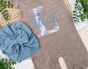 Può includere: Un completo per neonati composto da un pagliaccetto color beige con il nome "Milah" ricamato con un motivo floreale e una fascia blu chiaro con fiocco. Il pagliaccetto ha volant sulle maniche e sulle gambe.