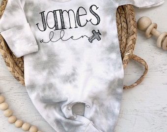 May include: A white and gray tie-dye baby onesie with long sleeves and a snap closure. The onesie has the name "James Allen" embroidered on the front, along with a small airplane.
