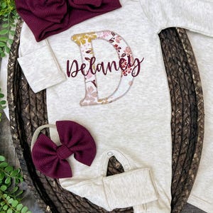 May include: A baby outfit set in cream color, featuring a long-sleeved onesie with the name "Delaney" and a floral initial "D". Includes a matching burgundy bow headband and a burgundy bow. The set is displayed on a woven tray.