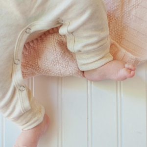 May include: A cream-colored long-sleeve onesie with metal snap closures along the inner leg seam, resting on a soft, pale pink knitted blanket with a textured diamond pattern. The garment features cuffed sleeves and legs for a snug fit.