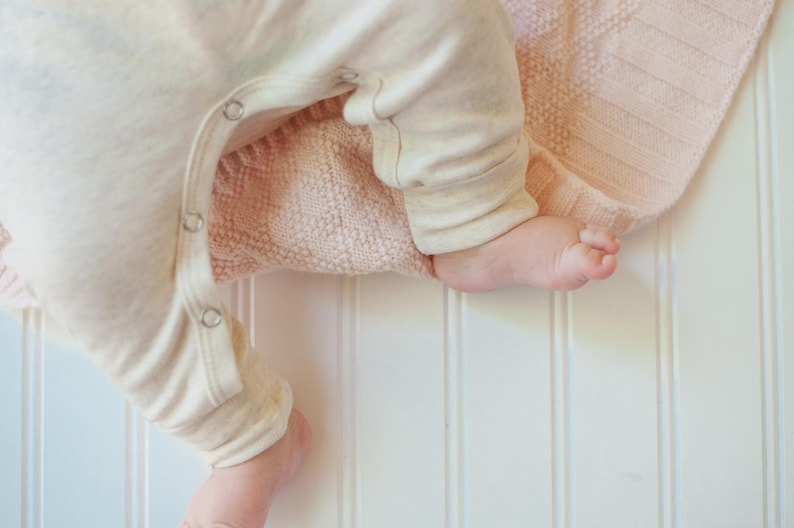 May include: A cream-colored long-sleeve onesie with metal snap closures along the inner leg seam, resting on a soft, pale pink knitted blanket with a textured diamond pattern. The garment features cuffed sleeves and legs for a snug fit.