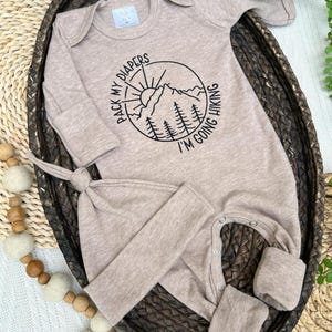 K&ouml;nnte beinhalten: Beige Baby-Schlafanzug und passendes Knotenm&uuml;tzchen-Set. Der Schlafanzug hat eine runde Grafik mit einer Berglandschaft und dem Text "PACK MY DIAPERS I'M GOING HIKING". Das Set ist aus weichem Material und f&uuml;r Neugeborene konzipiert.
