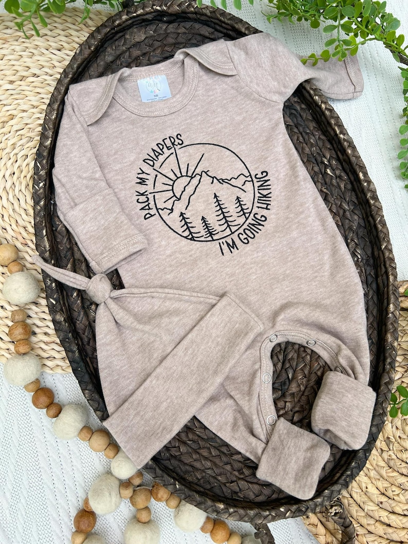 K&ouml;nnte beinhalten: Beige Baby-Schlafanzug und passendes Knotenm&uuml;tzchen-Set. Der Schlafanzug hat eine runde Grafik mit einer Berglandschaft und dem Text "PACK MY DIAPERS I'M GOING HIKING". Das Set ist aus weichem Material und f&uuml;r Neugeborene konzipiert.