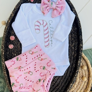 K&ouml;nnte beinhalten: Ein rosa-wei&szlig;es Baby-Outfit mit einem Zuckerstangen-Design. Das Outfit beinhaltet einen lang&auml;rmeligen Strampler mit dem Namen "Calliope" bestickt, ein Stirnband mit einer rosa Schleife und rosa-wei&szlig;e gemusterte Bl&uuml;mchen.