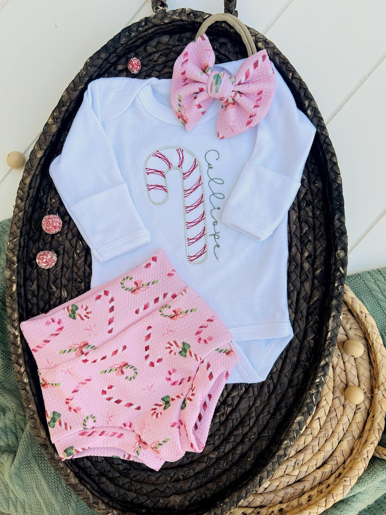 K&ouml;nnte beinhalten: Ein rosa-wei&szlig;es Baby-Outfit mit einem Zuckerstangen-Design. Das Outfit beinhaltet einen lang&auml;rmeligen Strampler mit dem Namen "Calliope" bestickt, ein Stirnband mit einer rosa Schleife und rosa-wei&szlig;e gemusterte Bl&uuml;mchen.