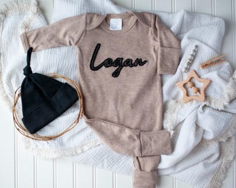 May include: A beige baby onesie with the name "Logan" embroidered in black on the front. The onesie is laid out on a white blanket with a black beanie and a wooden star rattle.