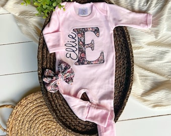 May include: Pink baby onesie with a floral letter E and the name "Ellie" embroidered on the front. The onesie is in a wicker basket with a floral bow.