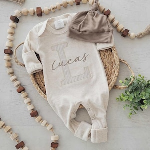 Könnte beinhalten: Ein langärmeliger cremefarbener Baby-Strampler mit einem großen gestickten Buchstaben L und dem Namen Lucas in Schreibschrift in der Mitte. Der Strampler wird mit einer passenden gerippten braunen Mütze mit Knoten geliefert.