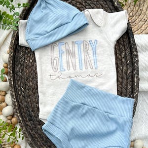 May include: A baby clothing set in a woven basket. The set includes a light blue ribbed knit hat, a short-sleeved onesie with the name "GENTRY" in blue and gray, and matching blue shorts. The name "thomas" is written below the name.