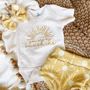 May include: A white baby onesie with a gold embroidered sun and the text "you are my sunshine". The onesie is paired with yellow shorts with a white sun pattern.