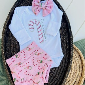 K&ouml;nnte beinhalten: Ein Baby-Outfit mit pink-wei&szlig;em Zuckerstangen-Muster, bestehend aus einem wei&szlig;en Langarm-Bodysuit, pink-wei&szlig;em Zuckerstangen-Muster-Bl&uuml;mchen und einem rosa Schleifen-Haarband. Der Bodysuit ist mit dem Namen "Calliope" bestickt.