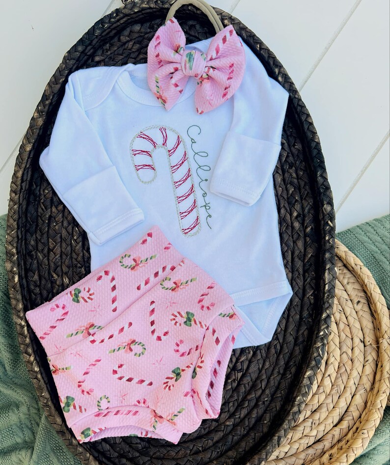K&ouml;nnte beinhalten: Ein Baby-Outfit mit pink-wei&szlig;em Zuckerstangen-Muster, bestehend aus einem wei&szlig;en Langarm-Bodysuit, pink-wei&szlig;em Zuckerstangen-Muster-Bl&uuml;mchen und einem rosa Schleifen-Haarband. Der Bodysuit ist mit dem Namen "Calliope" bestickt.