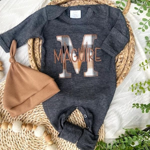 May include: A charcoal gray baby onesie with the name "MAGUIRE" embroidered in brown thread. The letter "M" is a plaid pattern in shades of brown, white, and tan. A matching brown knit cap is next to the onesie.