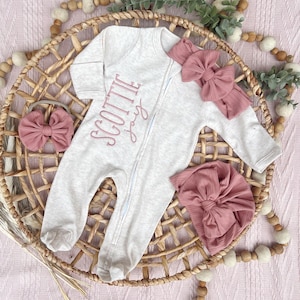 May include: A light gray baby sleeper with the name "SCOTTIE jay" embroidered in pink, a matching pink bow headband, a bow, and a knotted cap. The items are arranged in a woven basket on a pink surface.