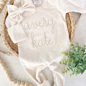 May include: A long-sleeve, cream-colored baby bodysuit featuring the embroidered name "avery kate" in a cursive font on the chest. The garment includes a matching ribbed fabric bow headband and has snap closures at the bottom for easy access.