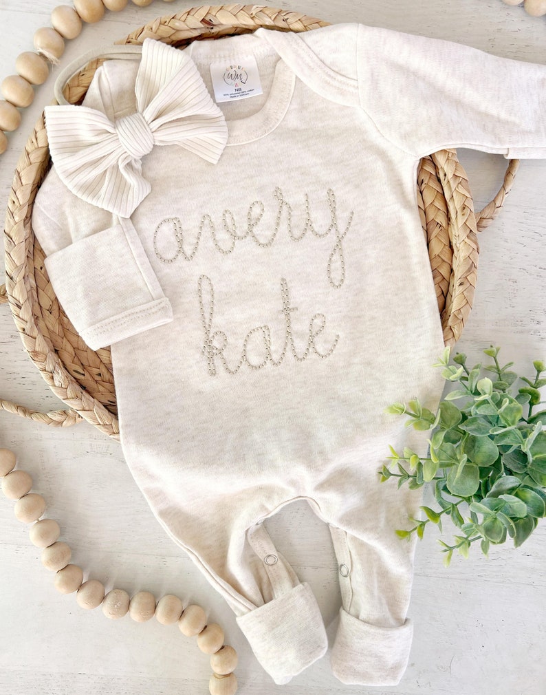 May include: A long-sleeve, cream-colored baby bodysuit featuring the embroidered name "avery kate" in a cursive font on the chest. The garment includes a matching ribbed fabric bow headband and has snap closures at the bottom for easy access.