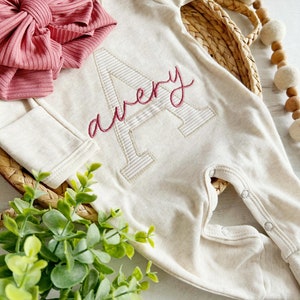 Puede incluir: Un pelele de bebé beige con mangas largas y una gran letra A bordada con el nombre "Avery" cosido en hilo rosa. Un lazo rosa está encima del pelele.