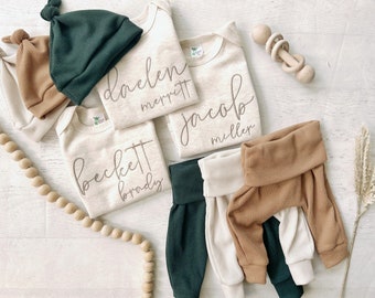 May include: Three beige baby onesies with personalised names, "Daelen Merritt", "Jacob Miller", and "Beckett Brady".  Two green and one beige ribbed knit baby hats.  One pair of green, one pair of beige, and one pair of brown ribbed knit baby trousers.  A wooden rattle.