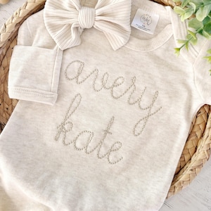 May include: A cream-colored long-sleeve baby bodysuit featuring the name Avery Kate embroidered in a light tan thread across the chest. A matching ribbed cream-colored bow headband is placed above the neckline of the garment.