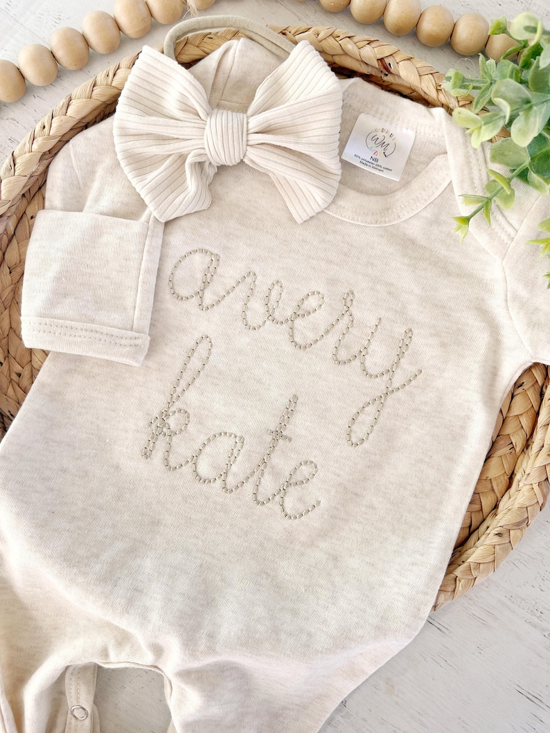 May include: A cream-colored long-sleeve baby bodysuit featuring the name Avery Kate embroidered in a light tan thread across the chest. A matching ribbed cream-colored bow headband is placed above the neckline of the garment.