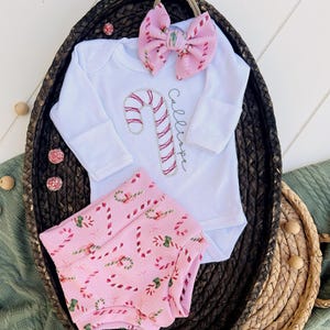 K&ouml;nnte beinhalten: Ein wei&szlig;er Langarm-Strampler mit einem Zuckerstangen-Design und dem Namen "Callie", der auf der Vorderseite gestickt ist. Der Strampler wird mit rosa und wei&szlig;en Zuckerstangen-Druck-Bl&uuml;mchen und einer rosa und wei&szlig;en Schleife kombiniert.