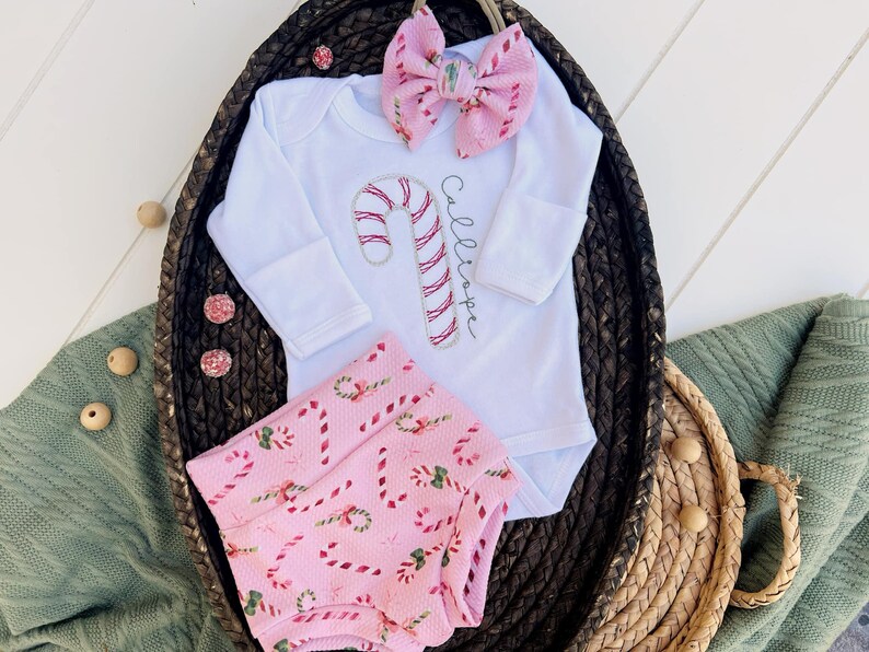 K&ouml;nnte beinhalten: Ein wei&szlig;er Langarm-Strampler mit einem Zuckerstangen-Design und dem Namen "Callie", der auf der Vorderseite gestickt ist. Der Strampler wird mit rosa und wei&szlig;en Zuckerstangen-Druck-Bl&uuml;mchen und einer rosa und wei&szlig;en Schleife kombiniert.