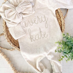 May include: A long-sleeved, cream-colored baby bodysuit featuring the embroidered name "avery kate" in a cursive font on the chest, paired with a matching cream-colored ribbed fabric headband adorned with a large, oversized bow detail.
