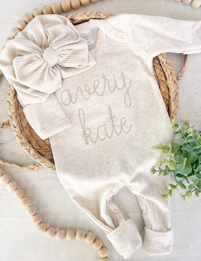 May include: A long-sleeved, cream-colored baby bodysuit featuring the embroidered name "avery kate" in a cursive font on the chest, paired with a matching cream-colored ribbed fabric headband adorned with a large, oversized bow detail.