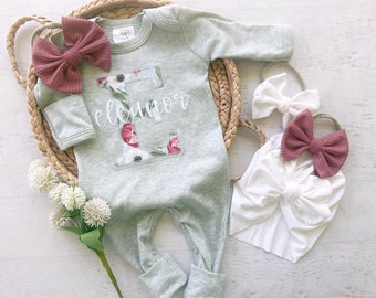 May include: A gray baby onesie with a floral letter E and the name "eleanor" embroidered on the front. The onesie is paired with two headbands, one white and one pink.